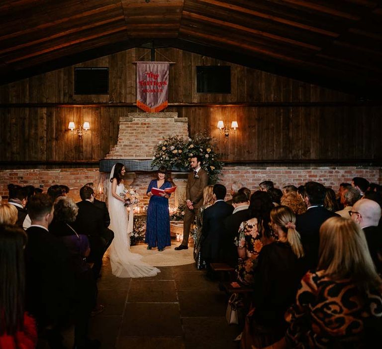 Groom in chocolate brown grooms suit standing with bride in strapless ivory fringed crepe gown and chapel length veil and celebrant in deep blue playsuit standing by large fireplace at Willow Marsh Farm Loughborough 