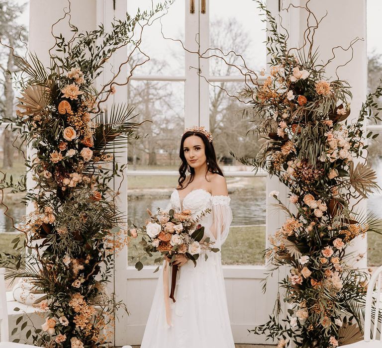 Bride in off the shoulder sheer sleeves holding wedding bouquet in front of pretty wedding flower columns 