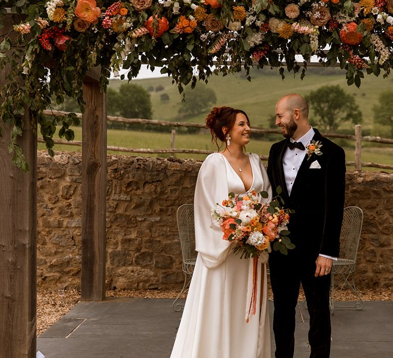 Bride in satin balloon sleeve Watters wedding dress posing with groom in traditional black tuxedo 