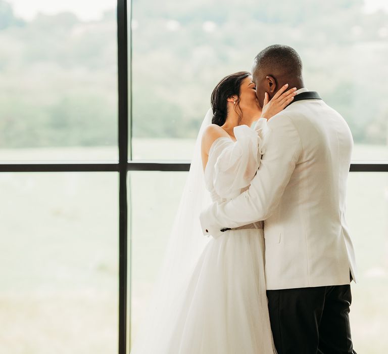 Groom in black and white tuxedo kisses bride in white tulle wedding dress in front of large window at Crumplebury wedding