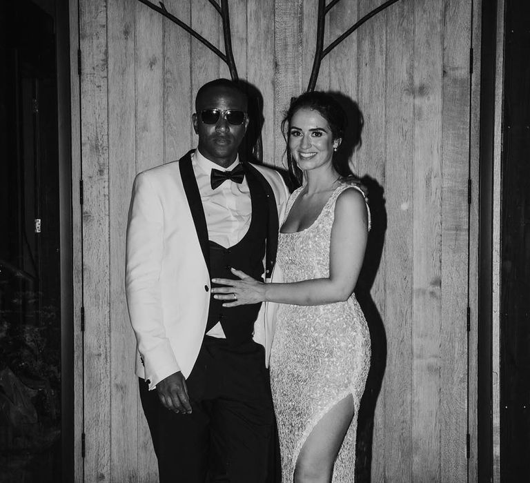 Groom in black-tie tuxedo and sunglasses poses with bride in fitted sparkly wedding reception dress