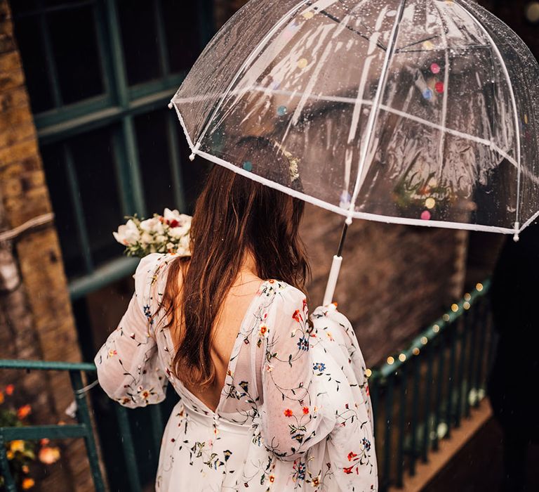 Bride holding clear umbrella wearing a Sassi Holford wedding dress with floral embroidery 