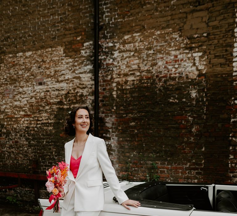 Bride wearing white wedding suit posing against the retro wedding car holding hot pink dried flower wedding bouquet 