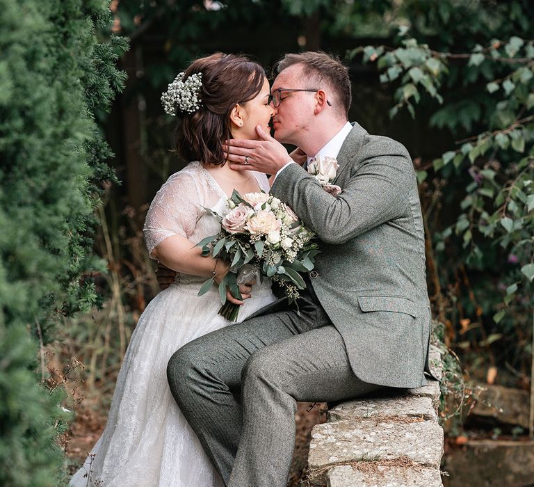 The groom in a grey suit sits with the bride in a lace wedding dress as they kiss for their couple portrait 