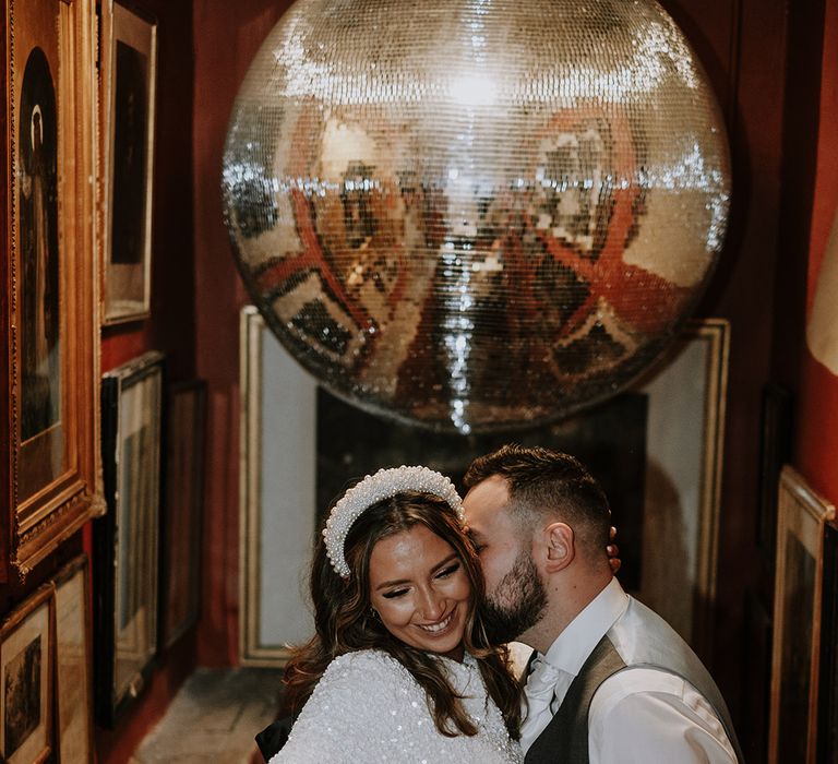 Bride wears long sleeve short wedding reception dress covered in white sequins with pearl headband with the groom 