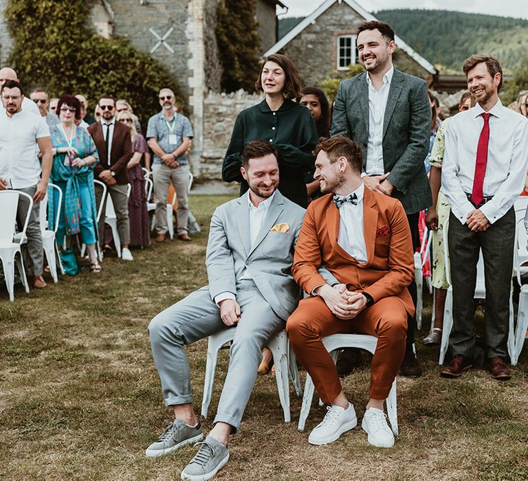 Wedding guests stand at Wilde Lodge wedding ceremony with grooms seated together 