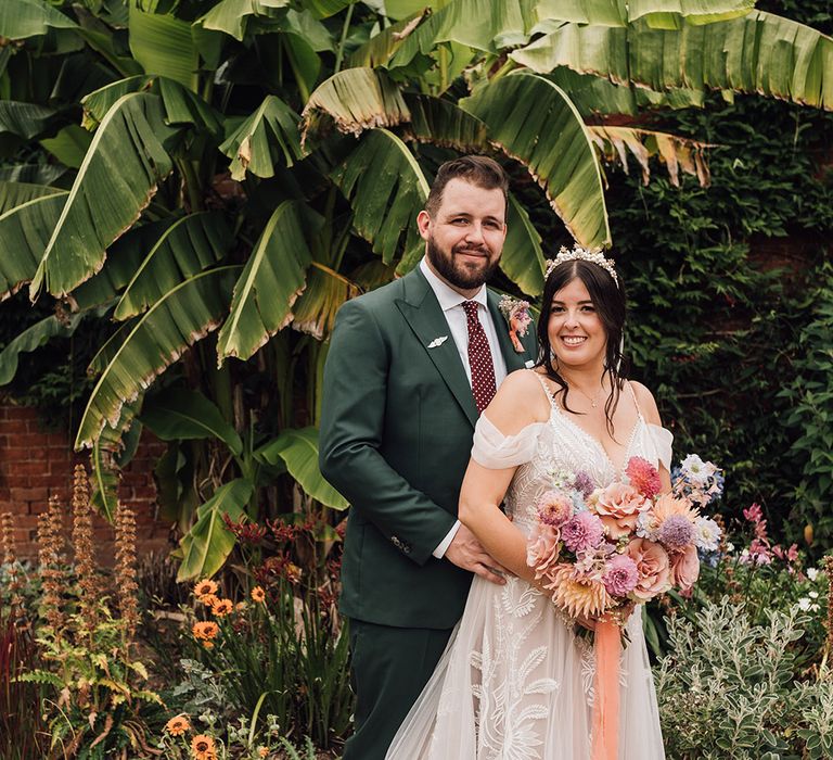 Bride in lace wedding dress with colourful bouquet with groom in green wedding suit 