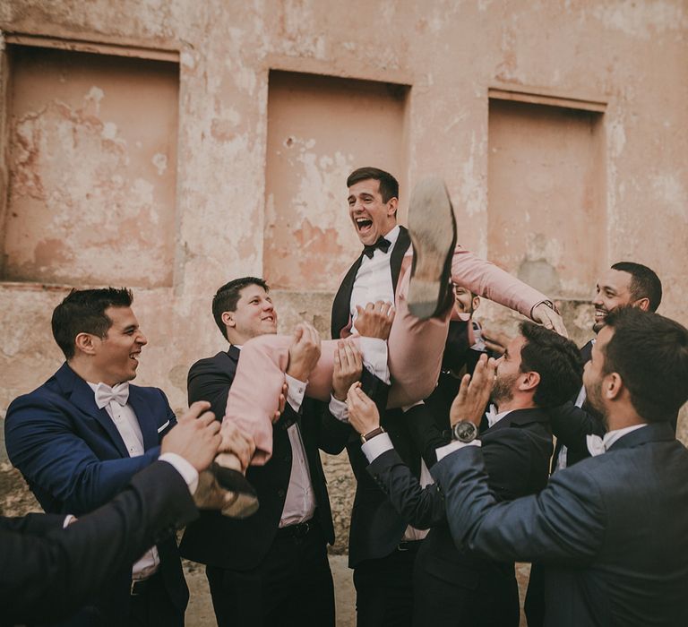 Groomsmen holding the groom in a pink tuxedo up in the air 