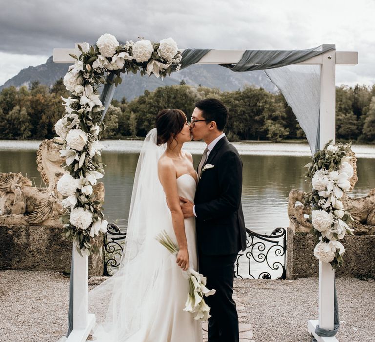 Bride and groom kissing next to a wooden frame decorated with drapes and white flowers 