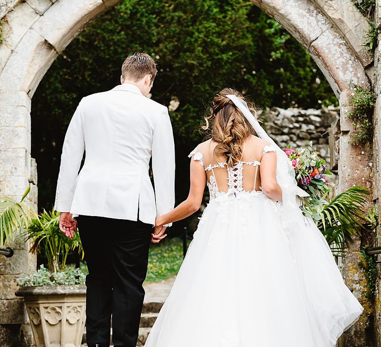 Bride with half up half down hair and cathedral length veil walking up the steps with her groom in a Galia Lahav wedding dress with illusion back