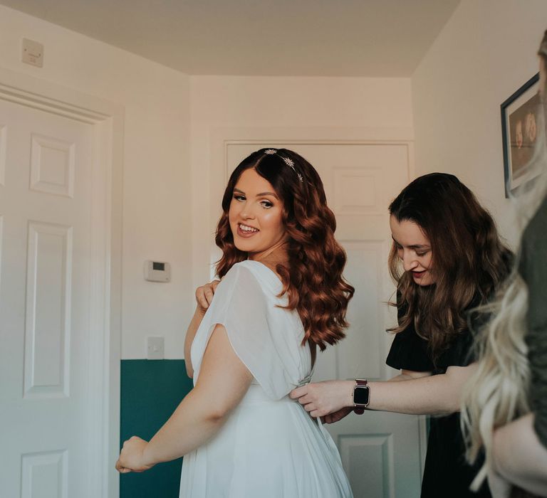 Bride with glam waves and celestial headband is zipped into white wedding dress