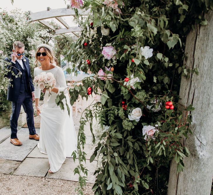 Smiling bride in Elbeth Gillis gown and sunglasses holding blush pink rose bouquet walks through Orangery at Euridge Manor with Groom in navy suit and sunglasses