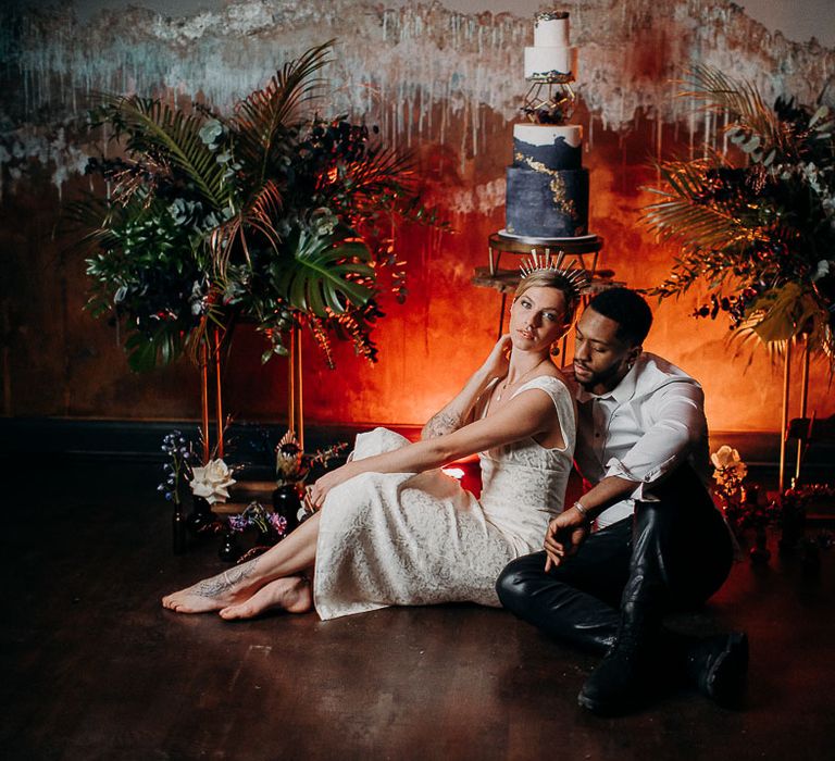 Stylish bride and groom sitting on the floor next to their black and white wedding cake flanked by moody dark wedding flowers arrangements 