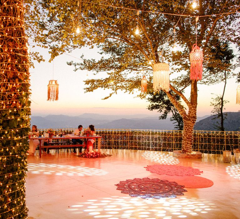 String lights covering the trees and railings, lanterns in the trees and guests sitting at the table as the sun begins to set