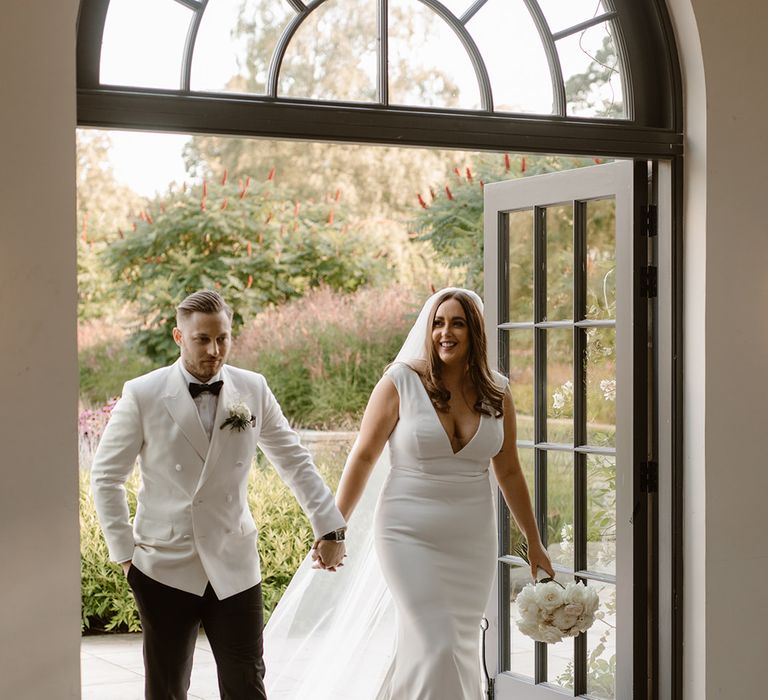 Bride in Made With Love wedding dress and groom in white double breasted tuxedo jacket enter the Fig House at Middleton Lodge holding hands