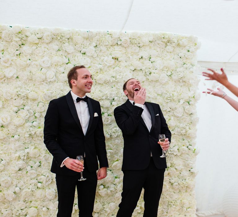 Grooms wearing black tuxedos stand by white flower wall