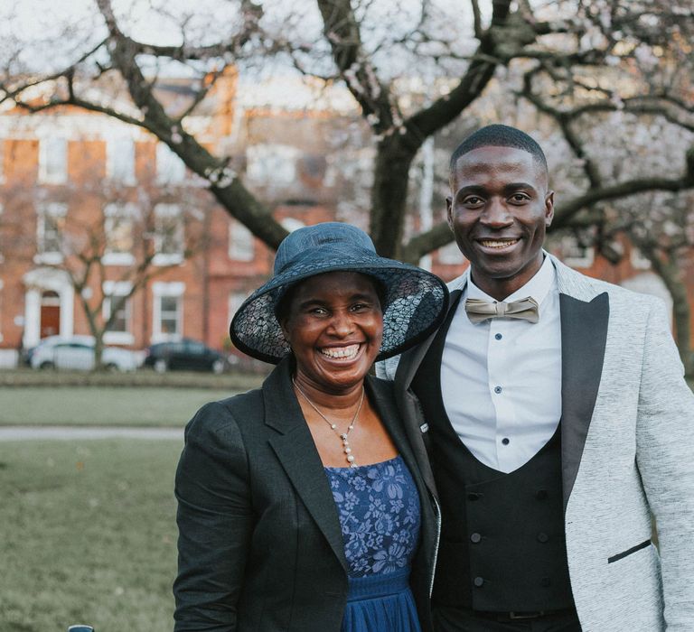 Groom in grey suit jacket with black lapels and gold bow tie stands smiling with wedding guests in blue dress, blue lace brimmed hat and black blazer outside after Bridge Community Church wedding