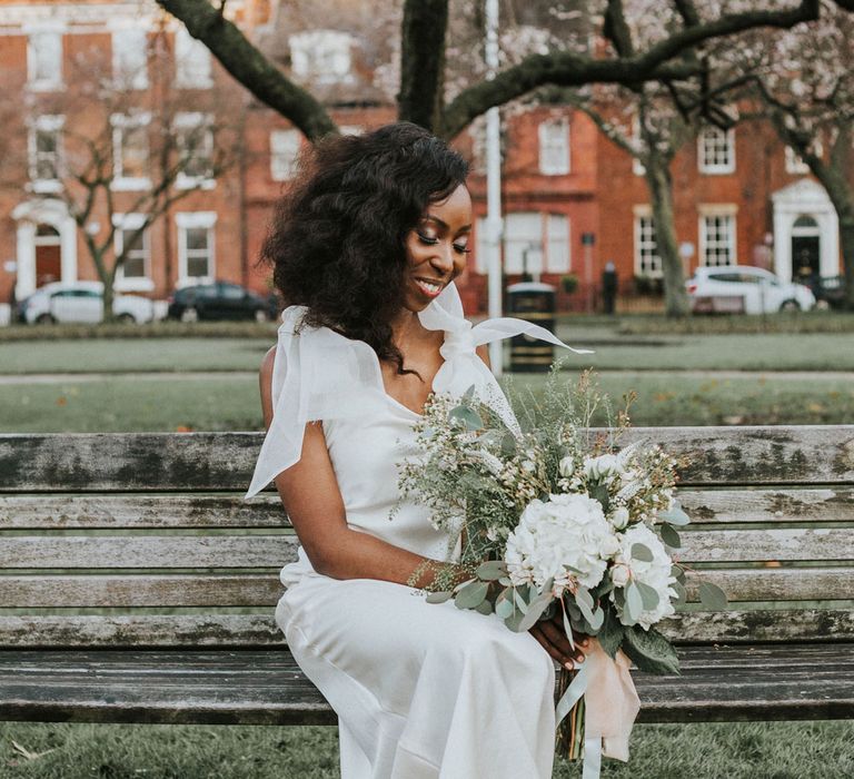 Bride in white satin Bec + Bridge wedding dress and silver heeled sandals sits on bench holding white and green bouquet after Bridge Community Church wedding