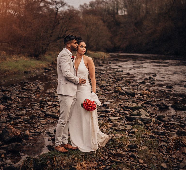 Bride & groom kiss outdoors as bride holds red floral bouquet 