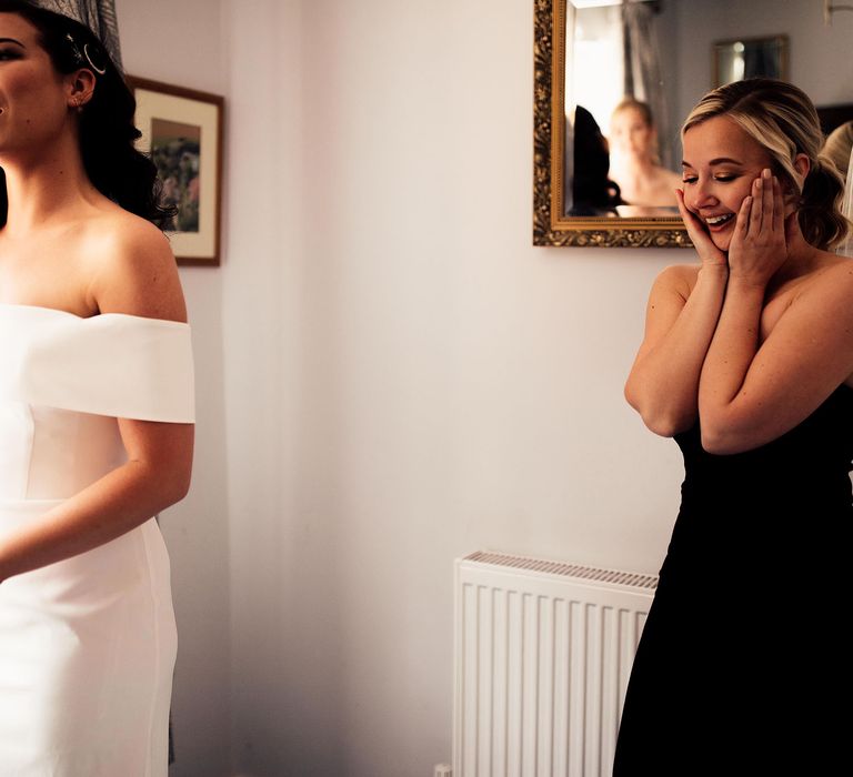 Lusher in strapless black dress holds face in her hands smiling at bride in strapless Vagabond wedding dress