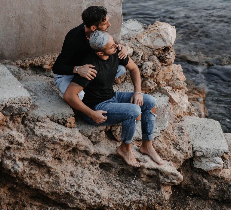 Grooms sit on rocks and look out to sea during engagement session