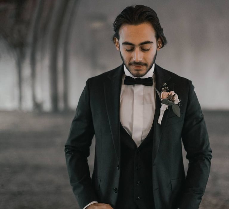 Groom looks down as he wears black tie attire with floral buttonhole 