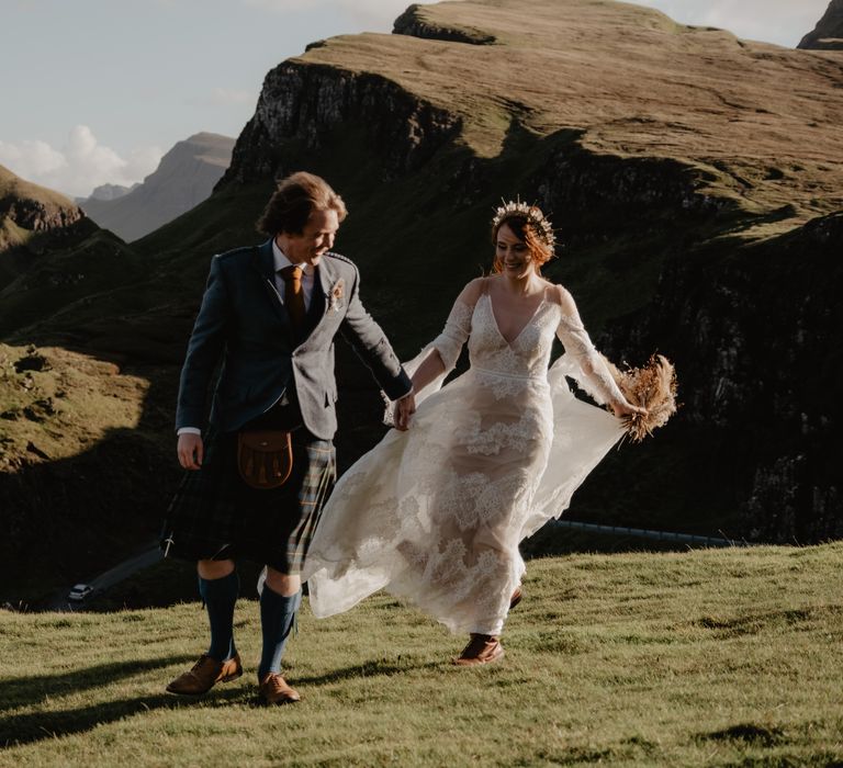 Bride lifts wedding gown and she walks along the hillside with her groom