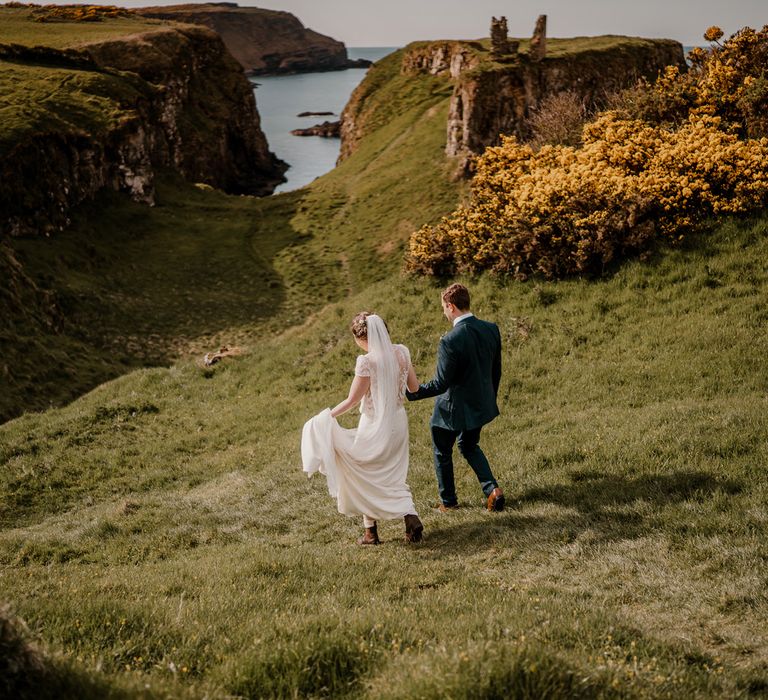 Smiling bride in lace top capped sleeve wedding dress with satin skirt holds hands of groom whilst they walk down hill after Dunluce Castle wedding