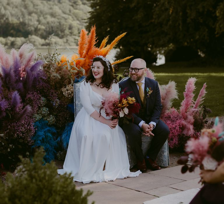 Bride & groom sit outdoors surrounded by colourful pampas grass in the countryside