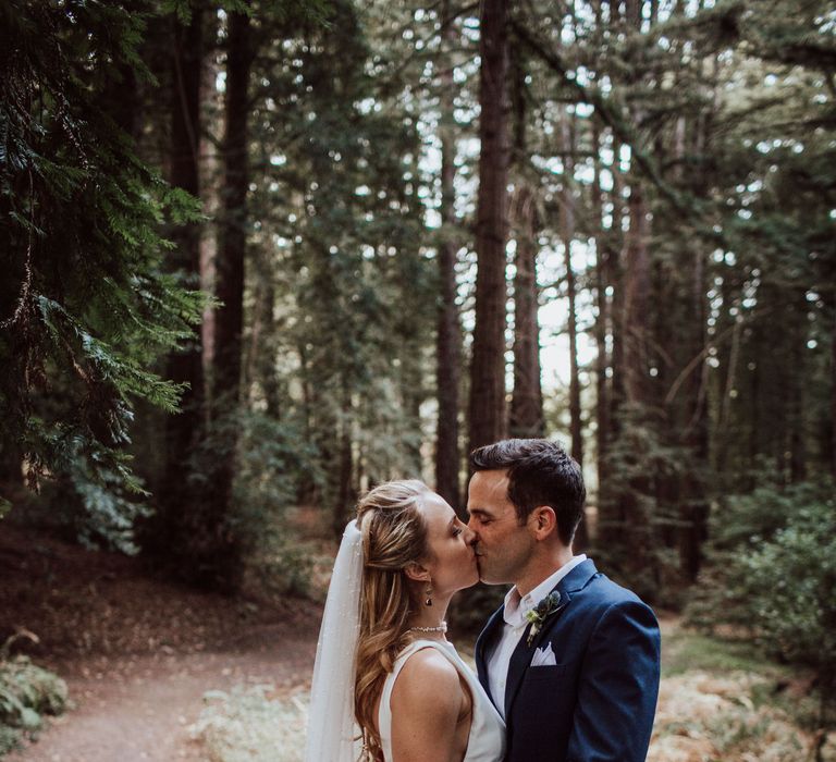 Bride & groom kiss in the woodland as the bride wears pearl wedding veil and holds peach bouquet