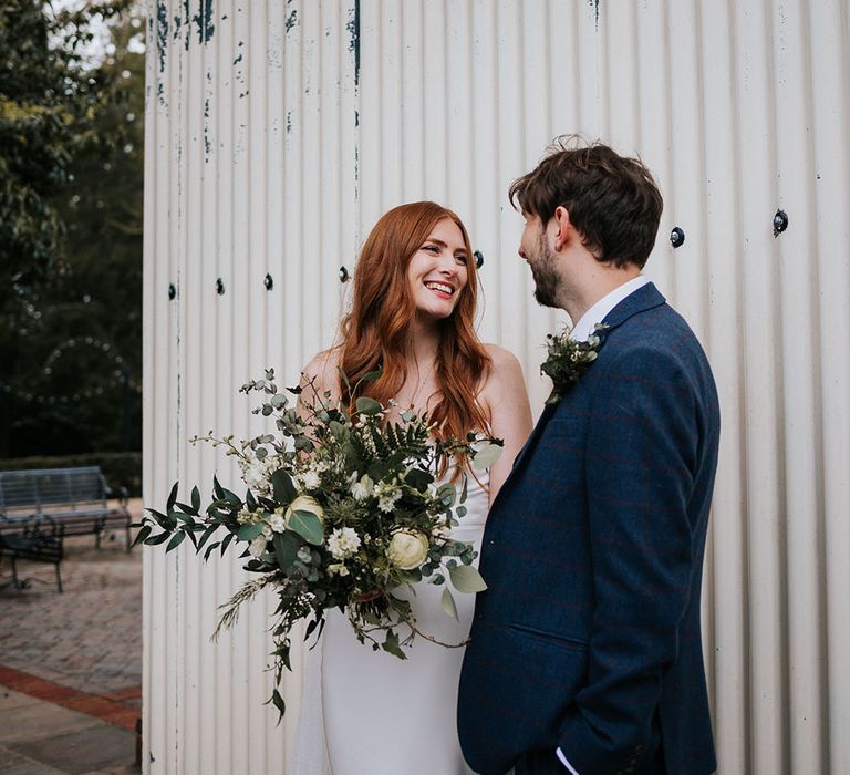 Beautiful bride holding a green foliage and white flower bouquet smiling at her husband in a navy suit 