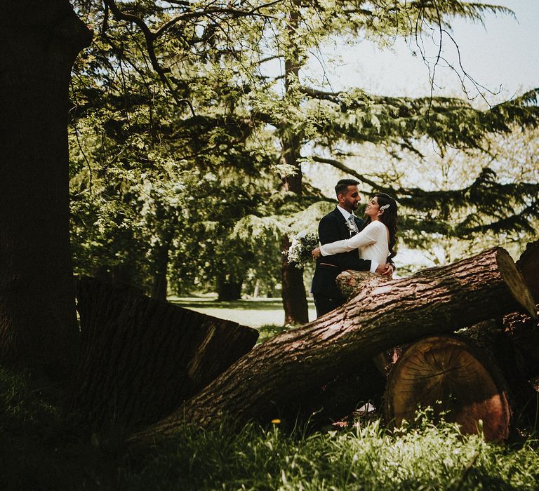 Groom in a navy blue suit embracing his bride in a long sleeve wedding dress by some logs 