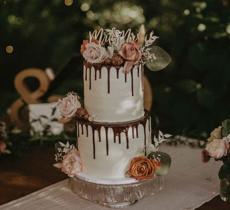 Two tier white wedding cake with chocolate drip detail, mixed burnt orange and pink roses and wooden topper reading Mr and Mrs for garden wedding with burnt orange wedding theme