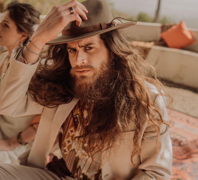 Groom wears his hair long and natural whilst lifting his hand to pick up his suede brown hat 