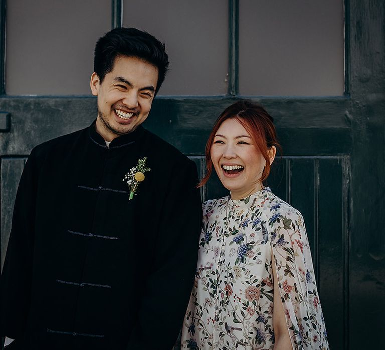 Portrait of an East Asian bride and groom smiling, the groom wears a navy velvet jacket and the bride a blue floral wedding dress with cape sleeves 