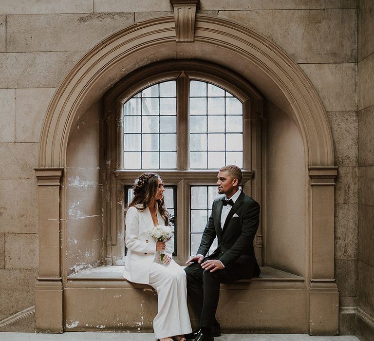 Groom in a tuxedo and bride in a white suit sitting on a window ledge at their Sheffield registry office wedding 