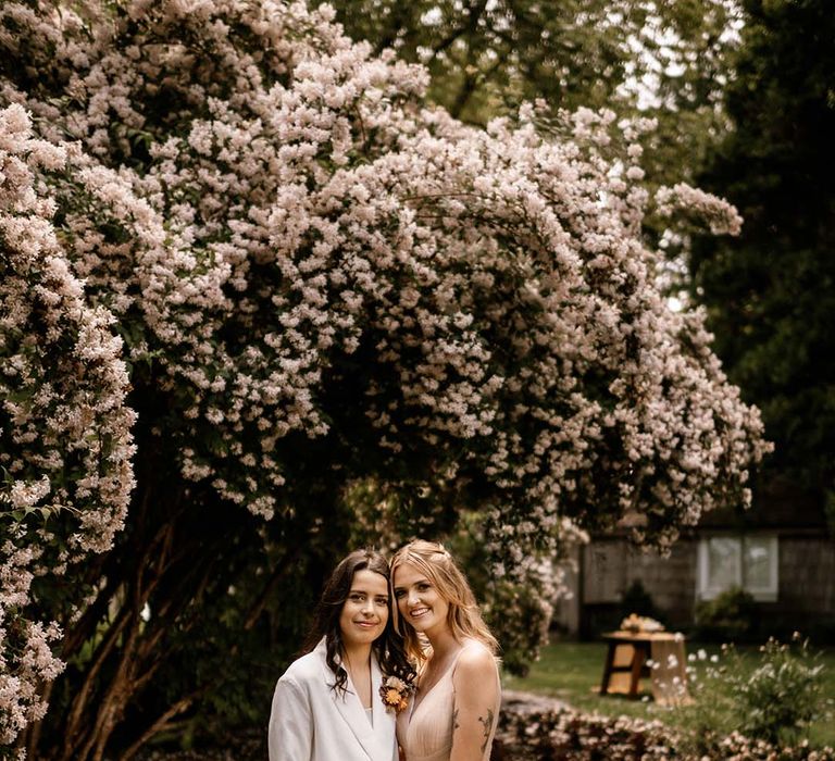 Two brides in a white wedding suit and blush wedding dress embracing under a blossom tree