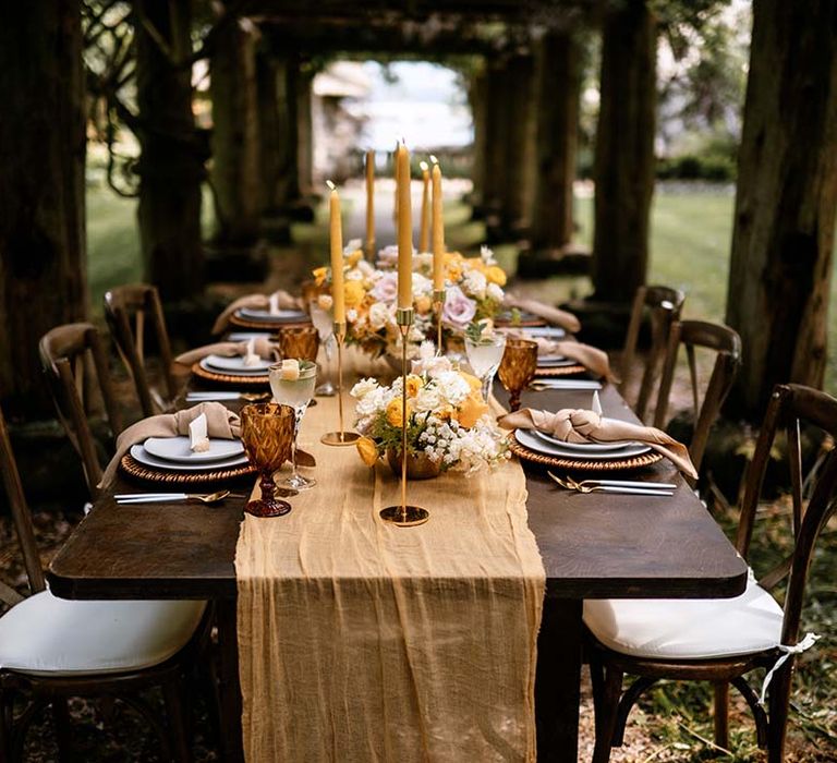 Alfresco wedding reception table decor with wooden table, yellow table runner and gold candlesticks 