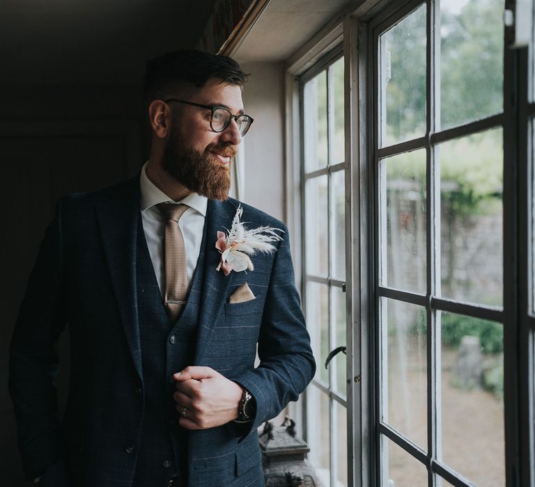 Groom looks out window as he wears three piece suit with floral buttonhole on his wedding day