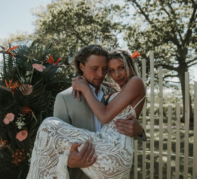 Groom in linen suit holds bride in white lace boho wedding dress during outdoor wedding ceremony with tropical flowers for birds of paradise wedding inspiration