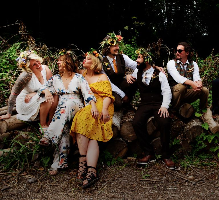 Grooms sit with their wedding guests on log outdoors in the woodland for midsummer vibe wedding 