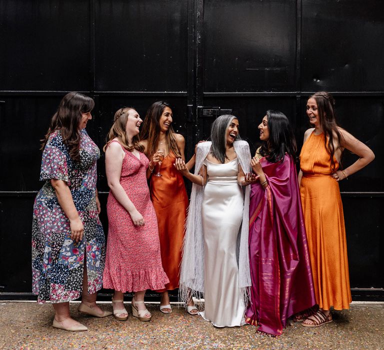 Bride in satin Halfpenny London wedding dress with tasselled bridal cape stands with wedding guests in multicoloured dresses outside at Loft Studios London