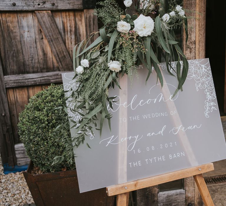 Frosted acrylic wedding welcome sign with leaf details topped with white floral decor at Tythe Barn wedding with barn wedding flowers