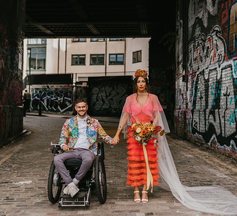 C5/C6 Tetraplegic groom in a wheelchair holding hands with his bride in a coral wedding dress and cape veil at their Shoreditch elopement 