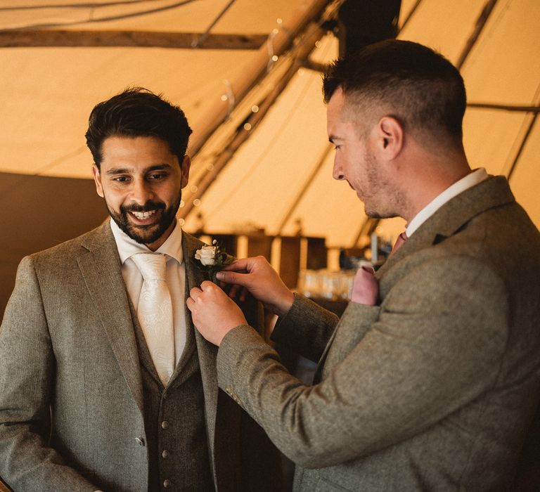 Groom in grey three piece suit with white tie has his floral buttonhole adjusted by groomsman in grey suit with pink pocket square inside tipi at Inkersall Grange Farm