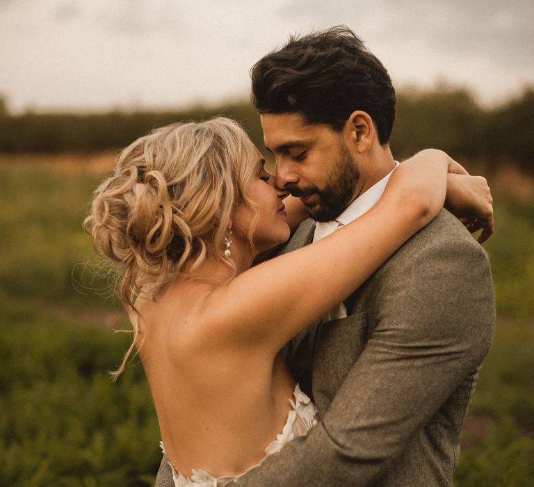 Bride in off the shoulder Enzoani wedding dress with curled wedding updo hugs groom in grey suit as they stand in field at Inkersall Grange Farm wedding