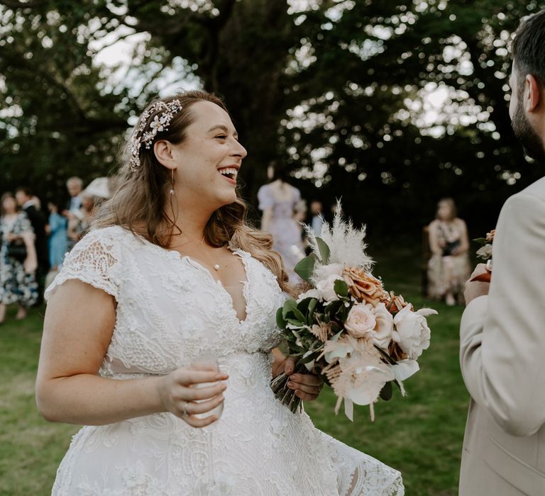 Birde in homemade lace wedding dress with pearl and floral hair pin holding mixed rose bouquet smiles at groom in beige suit at outdoor wedding reception