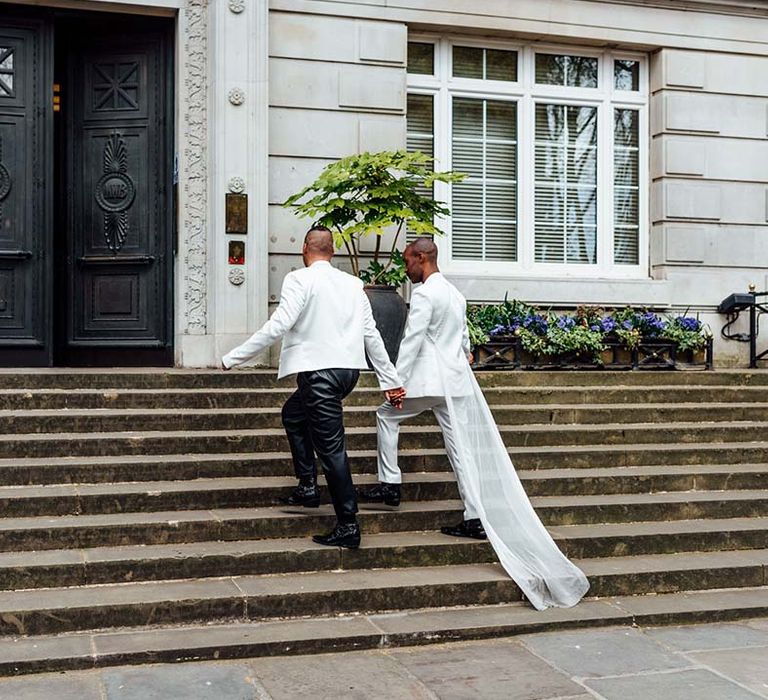 two stylish grooms in a white satin suit with a cape, leather trousers and double breasted jacket walking up the steps 