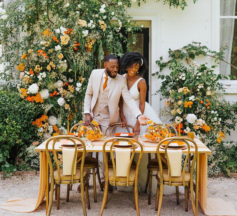 Black bride and groom in a beige suit and plunging neckline wedding dress at their outdoor wedding reception with orange decor and flowers 