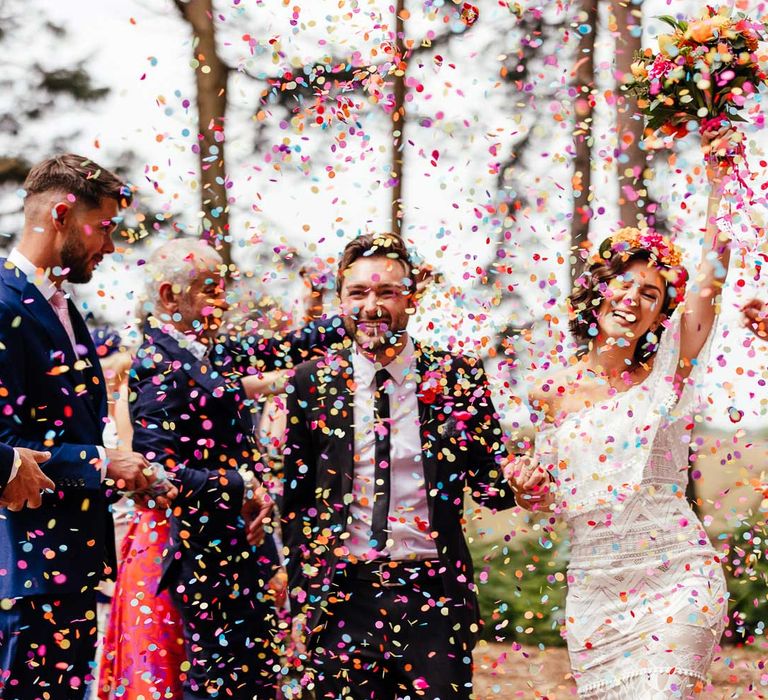 Bride in off the shoulder Grace Loves Lace wedding dress and bridal flower crown holds colourful wedding bouquet and the hand of groom in black suit as guests throw multicoloured confetti during outdoor ceremony at The Bridal Barn wedding venue